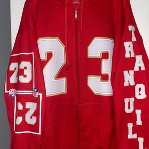 Puede incluir: Una sudadera roja con cremallera con el n&uacute;mero 23 en blanco y ribete dorado en la parte delantera y la manga izquierda. La manga derecha tiene la palabra "TRANQUIL" en blanco. La sudadera tiene capucha y cremallera completa.