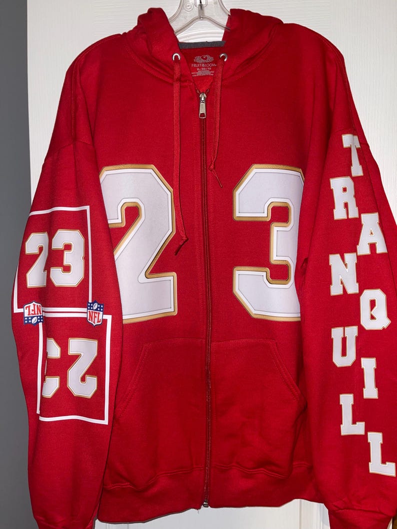 Puede incluir: Una sudadera roja con cremallera con el n&uacute;mero 23 en blanco y ribete dorado en la parte delantera y la manga izquierda. La manga derecha tiene la palabra "TRANQUIL" en blanco. La sudadera tiene capucha y cremallera completa.