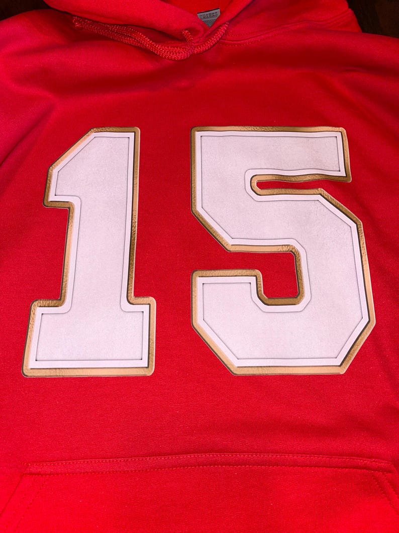 Puede incluir: Una sudadera con capucha roja con el n&uacute;mero "15" en la parte delantera. El n&uacute;mero es blanco con un contorno dorado y un borde blanco. La sudadera tiene un bolsillo delantero y una capucha.