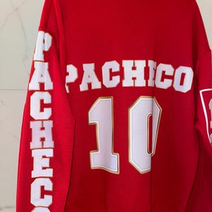 Puede incluir: Sudadera roja con el nombre "PACHICO" en letras blancas en la espalda y en la manga izquierda. El n&uacute;mero "10" est&aacute; debajo del nombre. Las letras y el n&uacute;mero tienen un contorno dorado.