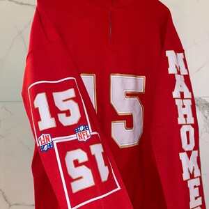 Puede incluir: Una sudadera roja con cuello con cremallera de un cuarto. El n&uacute;mero "15" se muestra prominentemente en blanco con ribete dorado en las mangas y la parte delantera. La palabra "MAHOMES" est&aacute; impresa verticalmente en la manga derecha.