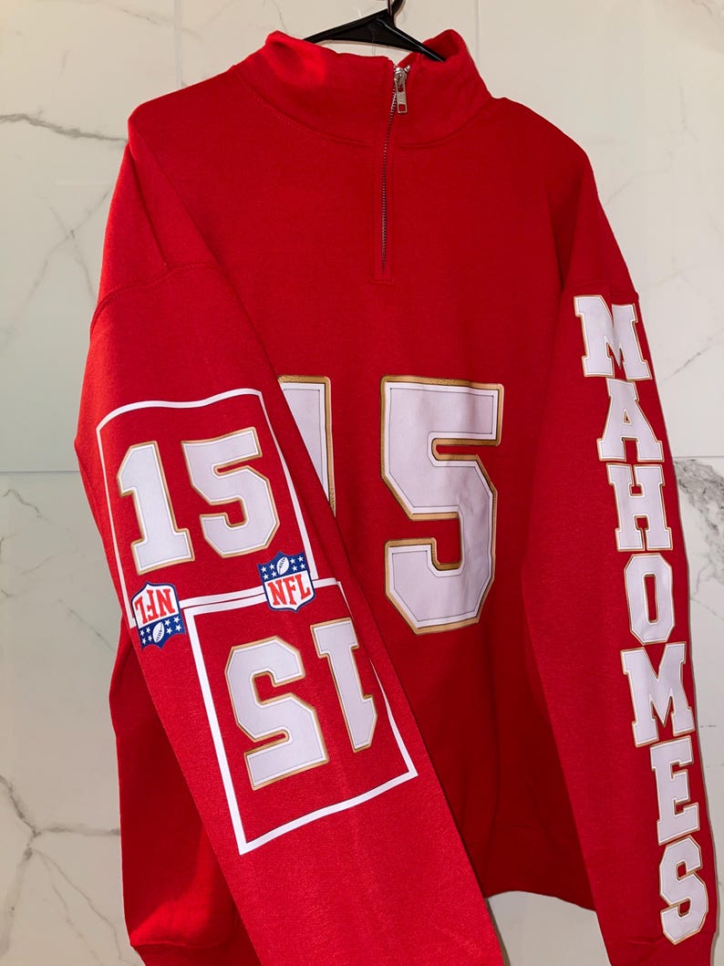 Puede incluir: Una sudadera roja con cuello con cremallera de un cuarto. El n&uacute;mero "15" se muestra prominentemente en blanco con ribete dorado en las mangas y la parte delantera. La palabra "MAHOMES" est&aacute; impresa verticalmente en la manga derecha.