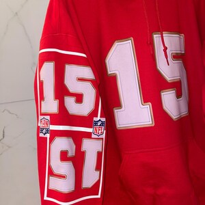 Puede incluir: Sudadera con capucha roja con el n&uacute;mero 15 en blanco con ribete dorado en la parte delantera y la manga. La manga tambi&eacute;n presenta dos logotipos de la NFL. La sudadera tiene un bolsillo delantero y una capucha con cord&oacute;n.