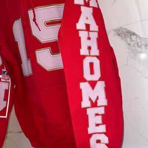 Puede incluir: Una sudadera roja con el n&uacute;mero 15 en blanco con ribete dorado en la parte delantera. La manga presenta el nombre "MAHOMES" en letras blancas con contornos rosas. Se ve un logotipo de la NFL.