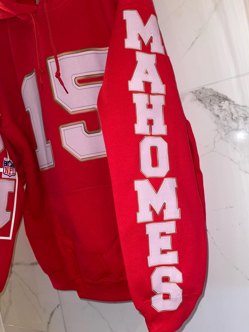 Puede incluir: Una sudadera roja con el n&uacute;mero 15 en blanco con ribete dorado en la parte delantera. La manga presenta el nombre "MAHOMES" en letras blancas con contornos rosas. Se ve un logotipo de la NFL.