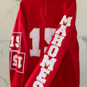 Puede incluir: Una sudadera roja con cuello alzado y cremallera. El n&uacute;mero "15" aparece en la parte delantera en blanco con contorno dorado. La manga derecha lleva el nombre "MAHOMES" en letras blancas. La manga izquierda tiene el n&uacute;mero "15" en un cuadrado.