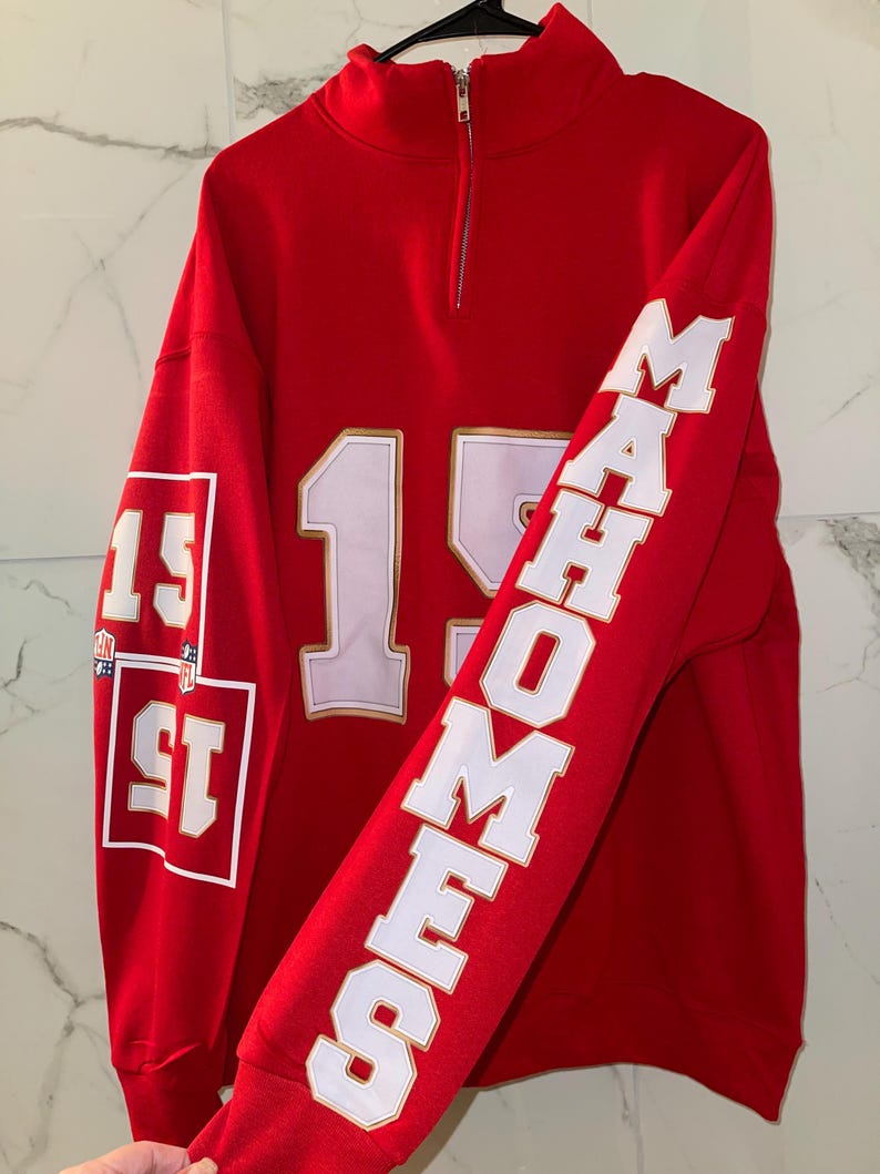 Puede incluir: Una sudadera roja con cuello alzado y cremallera. El n&uacute;mero "15" aparece en la parte delantera en blanco con contorno dorado. La manga derecha lleva el nombre "MAHOMES" en letras blancas. La manga izquierda tiene el n&uacute;mero "15" en un cuadrado.