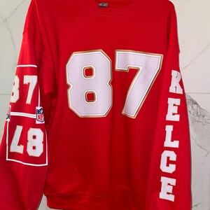 Puede incluir: Una sudadera roja con el n&uacute;mero 87 en blanco con un contorno dorado en la parte delantera. El nombre "KELCE" est&aacute; impreso verticalmente en la manga derecha en blanco. La manga izquierda tiene el n&uacute;mero 87 en un cuadro blanco.