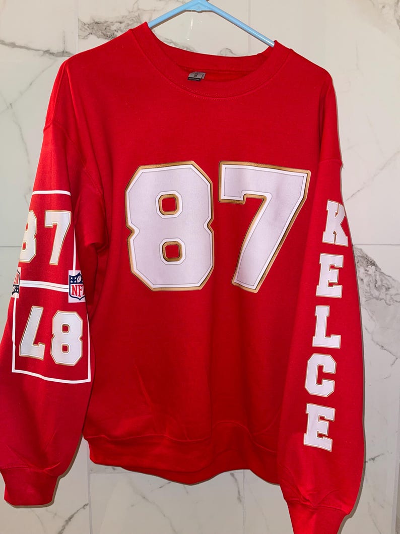 Puede incluir: Una sudadera roja con el n&uacute;mero 87 en blanco con un contorno dorado en la parte delantera. El nombre "KELCE" est&aacute; impreso verticalmente en la manga derecha en blanco. La manga izquierda tiene el n&uacute;mero 87 en un cuadro blanco.