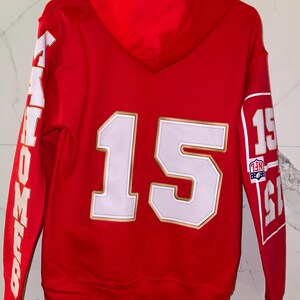 Puede incluir: Sudadera con capucha roja con detalles blancos y dorados. El n&uacute;mero 15 aparece en la espalda, con la palabra "MAHOMES" en la manga izquierda. La manga derecha muestra el n&uacute;mero 15 y un logotipo de la NFL.