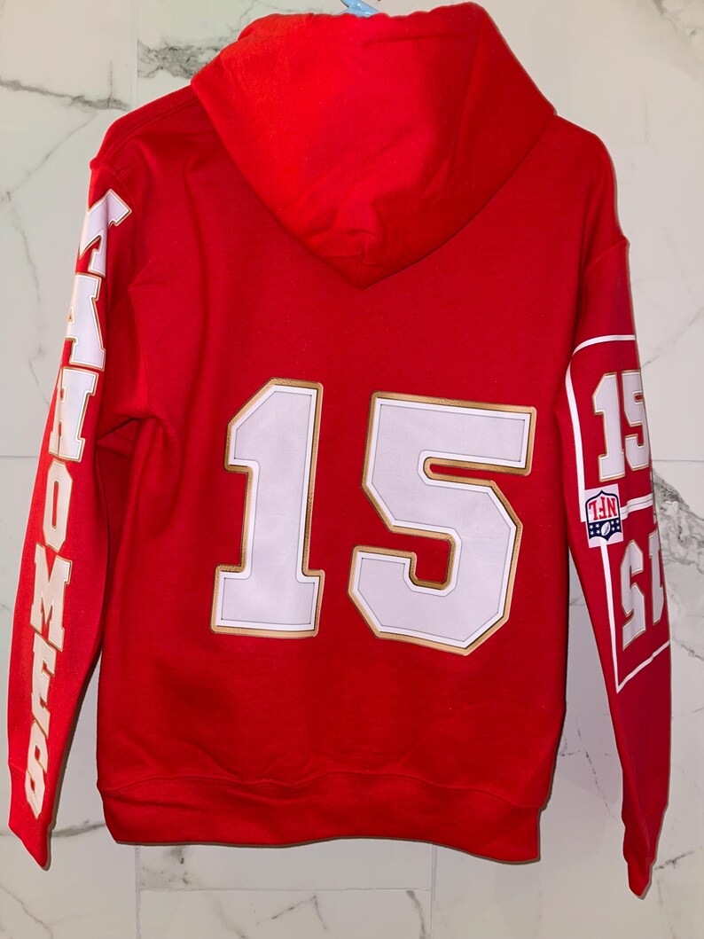 Puede incluir: Sudadera con capucha roja con detalles blancos y dorados. El n&uacute;mero 15 aparece en la espalda, con la palabra "MAHOMES" en la manga izquierda. La manga derecha muestra el n&uacute;mero 15 y un logotipo de la NFL.