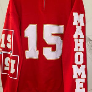 Puede incluir: Sudadera roja con detalles blancos y dorados. El n&uacute;mero 15 se muestra en letras blancas grandes con un contorno dorado en la parte delantera. La manga izquierda tiene el n&uacute;mero 15 en un cuadrado. La manga derecha dice "MAHOMES" en letras blancas.