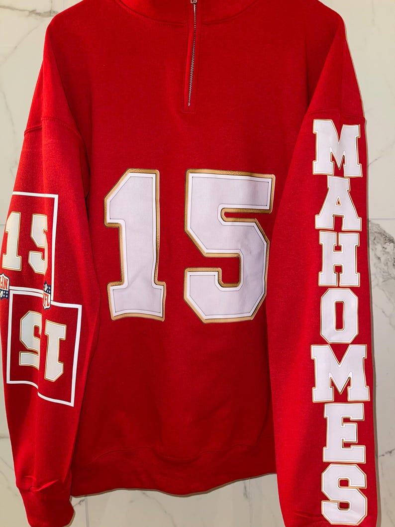 Puede incluir: Sudadera roja con detalles blancos y dorados. El n&uacute;mero 15 se muestra en letras blancas grandes con un contorno dorado en la parte delantera. La manga izquierda tiene el n&uacute;mero 15 en un cuadrado. La manga derecha dice "MAHOMES" en letras blancas.