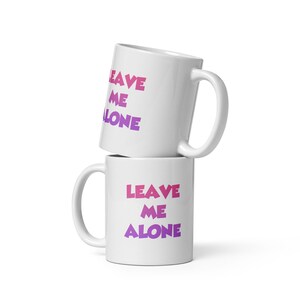 Puede incluir: Dos tazas de cerámica blanca apiladas una encima de la otra. La taza superior tiene el texto "LEAVE ME ALONE" en letras rosa y morado. La taza inferior tiene el mismo texto en una fuente más grande.