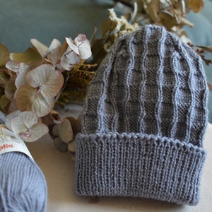 Könnte beinhalten: Eine graue gestrickte Baby-Mütze mit einem strukturierten Muster. Die Mütze ist aus Baby Nature Garn von Katia.