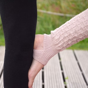 Könnte beinhalten: Hellrosa gestrickte Beinwärmer mit strukturiertem Muster. Die Beinwärmer bedecken Fuß und Wade, mit einem Ausschnitt für die Ferse. Die Person trägt eine schwarze Leggings. Der Hintergrund ist ein Holzdeck und grünes Gras.