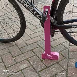 K&ouml;nnte beinhalten: Ein rosafarbener Fahrradst&auml;nder aus Metall mit einem Fahrrad-Silhouetten-Design. Der St&auml;nder dient dazu, ein Fahrrad zu sichern, indem das Hinterrad verriegelt wird.