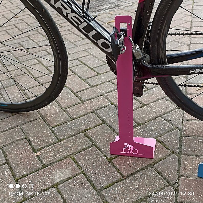 K&ouml;nnte beinhalten: Ein rosafarbener Fahrradst&auml;nder aus Metall mit einem Fahrrad-Silhouetten-Design. Der St&auml;nder dient dazu, ein Fahrrad zu sichern, indem das Hinterrad verriegelt wird.