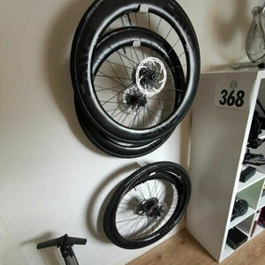 May include: Three black bicycle wheels with silver spokes and a black rim are mounted on a white wall. The wheels are stacked on top of each other. A black bicycle pump is on the floor below the wheels.