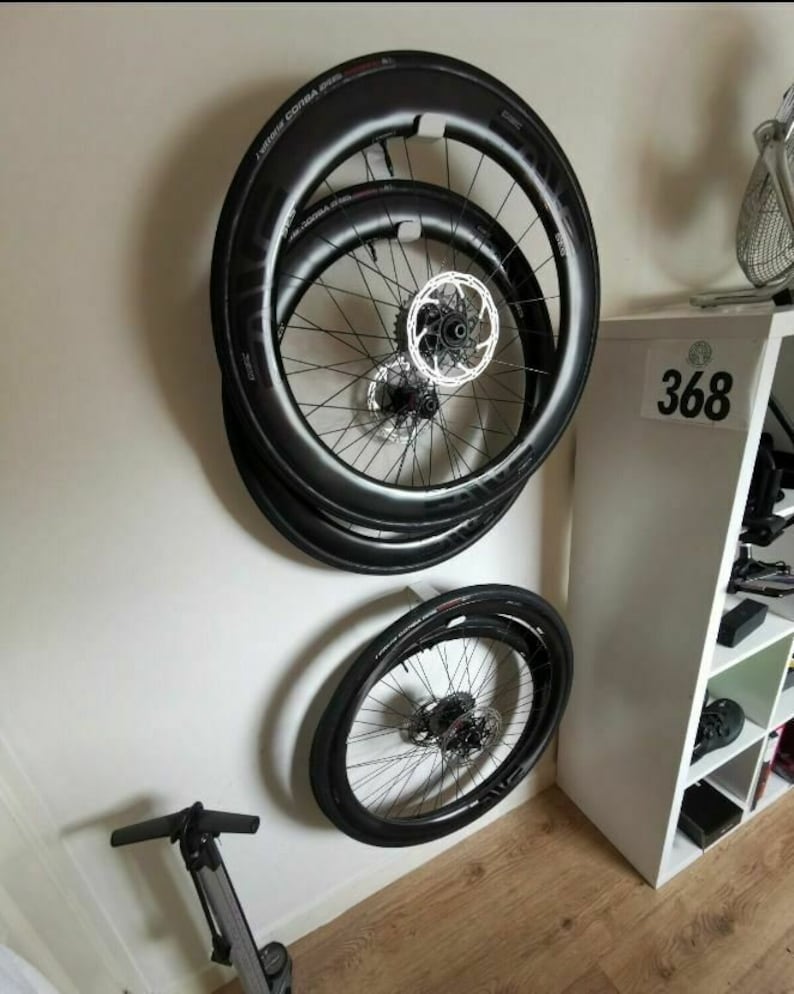 May include: Three black bicycle wheels with silver spokes and a black rim are mounted on a white wall. The wheels are stacked on top of each other. A black bicycle pump is on the floor below the wheels.