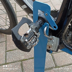 K&ouml;nnte beinhalten: Ein blauer Fahrradst&auml;nder aus Metall mit einem daran befestigten Fahrrad. Das Fahrrad hat einen schwarzen Rahmen und ein schwarzes und silbernes Pedal.