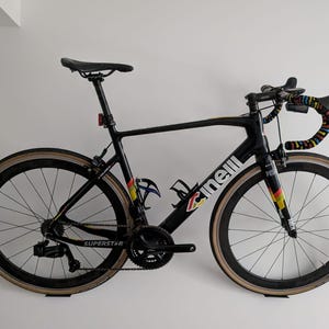 May include: A black Cinelli Superstar road bike with colourful handlebar tape and a black saddle. The bike has a black frame and black wheels with tan-coloured tyres.