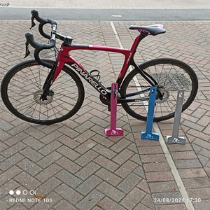 K&ouml;nnte beinhalten: Ein rosafarbenes Pinarello Prince Rennrad mit schwarzen Akzenten lehnt an drei Fahrradst&auml;ndern. Die St&auml;nder sind rosa, blau und grau.