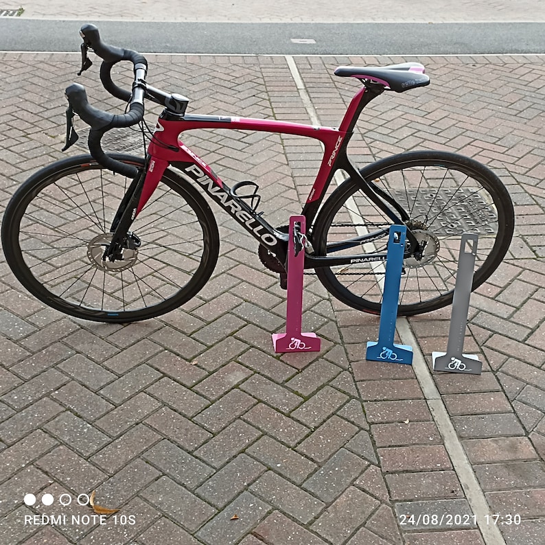 K&ouml;nnte beinhalten: Ein rosafarbenes Pinarello Prince Rennrad mit schwarzen Akzenten lehnt an drei Fahrradst&auml;ndern. Die St&auml;nder sind rosa, blau und grau.