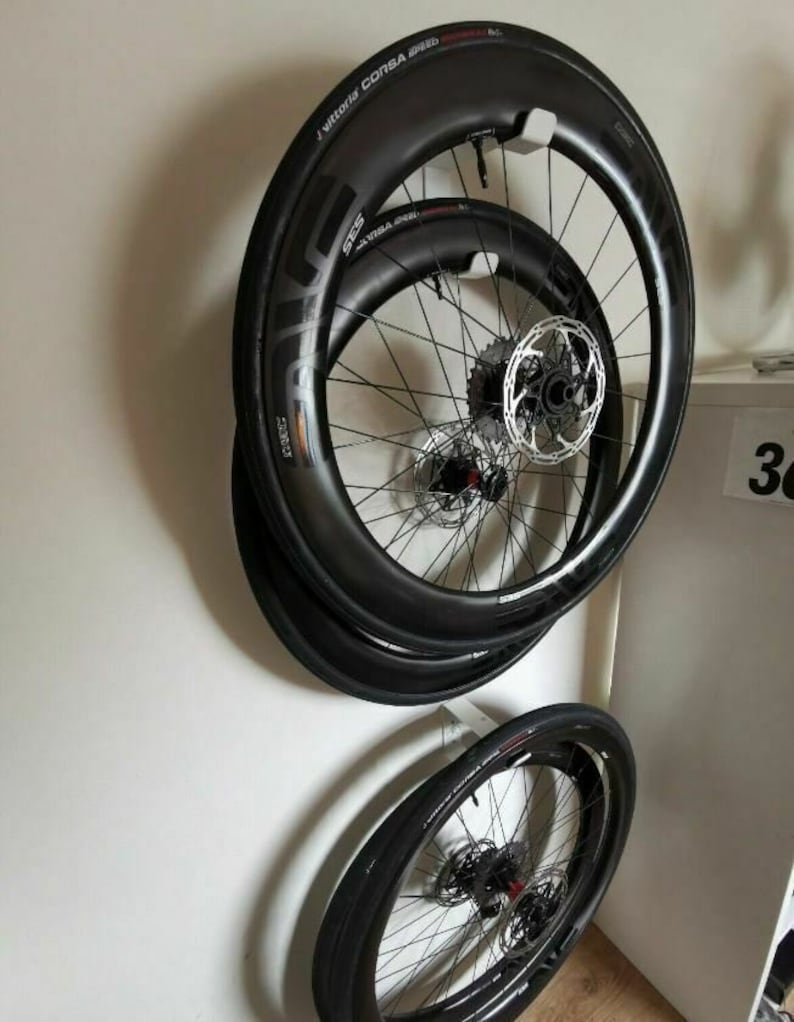 May include: Three black bicycle wheels with tires mounted on a white wall mount. The wheels are stacked on top of each other. The top wheel has the text "J Vittoria CORSA SPEED" printed on the tire.
