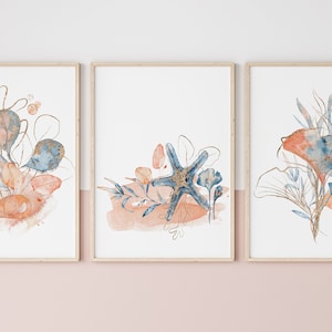 以下が含まれることがあります： 青とオレンジの色合いの抽象的な花柄が描かれた水彩画のプリント3枚セット。プリントは自然な木製のフレームに収められており、ミニマルな美しさがあります。