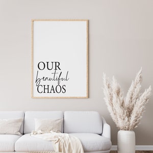 Puede incluir: Una impresión enmarcada con el texto "OUR beautiful CHAOS" en letras negras sobre fondo blanco. La impresión está colgada en una pared beige claro sobre un sofá blanco con una manta y almohadas. Un jarrón con hierba de la pampa seca está en el lado derecho de la imagen.