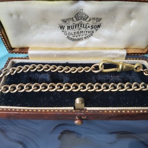 May include: A gold-toned metal watch chain with a swivel clasp, resting inside a rectangular brown leather presentation box. The interior lid features the printed text: ESTD 1850 W. RUFFELL & SON GOLDSMITHS TUNBRIDGE WELLS, set against cream fabric.