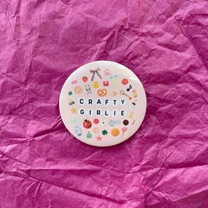Könnte beinhalten: Ein runder, hellrosa Button mit den Worten "CRAFTY GIRLIE" in schwarzen Blockbuchstaben. Der Button ist mit kleinen Illustrationen einer Schleife, einer Erdbeere, einer Brezel und anderen handwerklichen Elementen verziert. Der Button befindet sich auf einem rosa Hintergrund.
