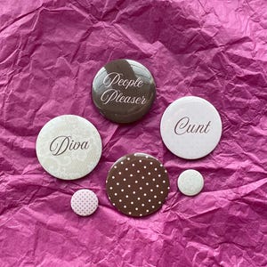 Könnte beinhalten: Eine Sammlung von sechs runden Buttons mit verschiedenen Designs und Texten. Die Buttons sind braun, weiß und rosa, mit Aufschriften wie "People Pleaser", "Diva" und "Cunt". Einige Buttons haben Polka Dots oder Spitzendesigns.