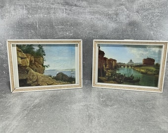 Op de afbeelding: Twee ingelijste landschapschilderijen. Het schilderij aan de linkerkant toont een rotsachtige klif met uitzicht op een waterlichaam. Het schilderij aan de rechterkant toont een stadsgezicht met een kanaal en een boot.