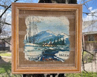 Könnte beinhalten: Ein gerahmtes Gemälde auf Jute, das eine Winterszene mit einem kleinen Dorf in einem verschneiten Tal darstellt. Das Gemälde ist in Blautönen, Weiß und Braun gehalten.