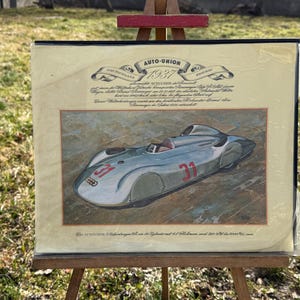 Puede incluir: Obra de arte enmarcada de un coche de carreras Auto Union vintage de 1937, expuesta sobre un caballete al aire libre. El coche es plateado con el número 31 en rojo. La obra incluye texto en alemán.