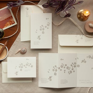 May include: A set of white wedding invitations with a delicate floral design in gray. The invitations are laid out on a table with a wooden surface and a beige linen tablecloth. There is a small seashell, a candle, and a vintage photograph on the table.