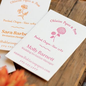 May include: Two white rectangular business cards with textured paper. One card features orange text and a flower illustration, while the other displays pink text and a hydrangea graphic. Both cards include the text Oblation Papers & Press.