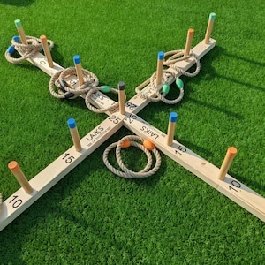 Op de afbeelding: Een houten ringwerpspel op een groene ondergrond. De set bevat een houten basis met genummerde pinnen en touwringen met gekleurde accenten. De pinnen zijn geschilderd in blauw, oranje en groen.