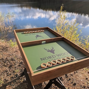 Pode incluir: Um jogo de mesa de madeira com uma superfície de jogo verde e o texto "NORD WOOD CREATIONS" e um logotipo de cabeça de veado. O jogo inclui discos de madeira e está montado ao ar livre perto de um lago.