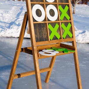 Könnte beinhalten: Ein hölzernes Tic-Tac-Toe-Spiel mit einer dunkelbraunen Spielfläche und einem hellbraunen Holzrahmen. Das Spiel hat weiße Os und grüne Xs. Das Spiel ist im Freien auf einer hellblauen Oberfläche aufgebaut.