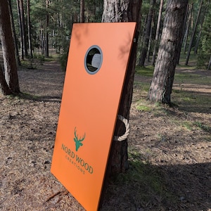Könnte beinhalten: Ein orangefarbenes Cornhole-Spielbrett mit einem kreisförmigen Loch und einem grünen Logo mit der Aufschrift "Nord Wood Creations". Das Brett lehnt an einem Baum in einer Waldumgebung. Ein Seilgriff ist befestigt.