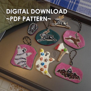 May include: A collection of handmade Halloween ornaments in various shapes, including ghosts, bats, and a coffin. The ornaments are made with cross-stitch and embroidery, featuring a variety of colors and designs. The text "DIGITAL DOWNLOAD ~PDF PATTERN~" is visible.