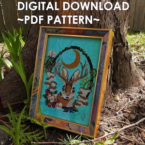 Puede incluir: Un patrón de punto de cruz enmarcado que presenta un conejo con cuernos, una luna creciente y elementos florales. La obra de arte está sobre un fondo turquesa y enmarcada en un marco dorado. El texto "DIGITAL DOWNLOAD ~PDF PATTERN~" se muestra en la parte superior.