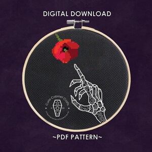 May include: A black and white cross stitch pattern featuring a skeletal hand reaching for a red poppy flower. The pattern is framed in a wooden embroidery hoop. The text "Curio Cabinet Crafts" is visible in the lower left corner of the pattern.