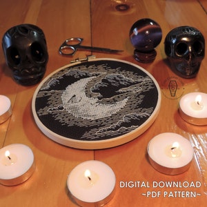 May include: A black and white cross stitch pattern of a crescent moon in the clouds. The pattern is in a wooden embroidery hoop on a wooden table with candles and other decorative items.