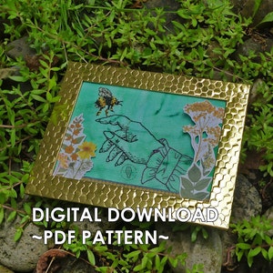 May include: A cross-stitch pattern featuring a hand reaching out to a bee, surrounded by floral elements. The pattern is framed in a gold frame with a honeycomb design. The text "DIGITAL DOWNLOAD - PDF PATTERN -" is displayed below the frame.