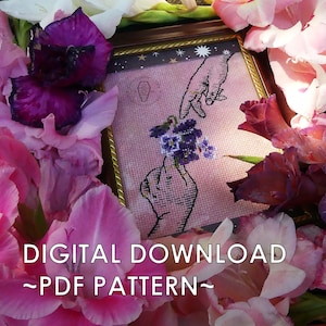 May include: Cross stitch pattern featuring two hands reaching towards each other, with a cluster of purple flowers on one finger. The pattern is framed and surrounded by pink and purple flowers.