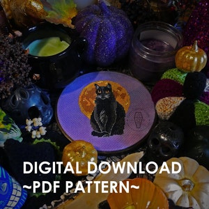 Puede incluir: Un patrón de punto de cruz con un gato negro sentado frente a una luna llena. El patrón se muestra en un aro de bordado redondo rodeado de decoraciones de Halloween, incluyendo calabazas, calabacines y velas. El texto "DIGITAL DOWNLOAD ~PDF PATTERN~" se muestra en la parte inferior de la imagen.